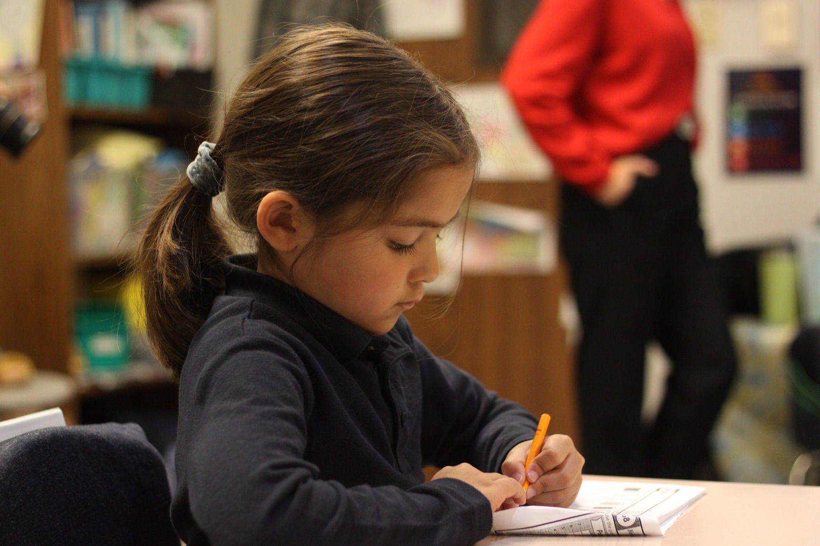 A NOCA Lower School student writing at her desk, focused on her work, a teacher standing by.