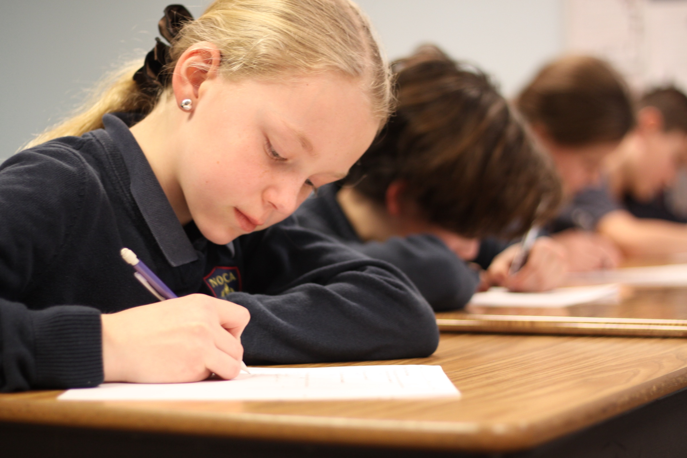 A NOCA student writes at her desk with a pencil, the NOCA crest visible on her navy sweater. A row of classmates behind her are working at the same task.