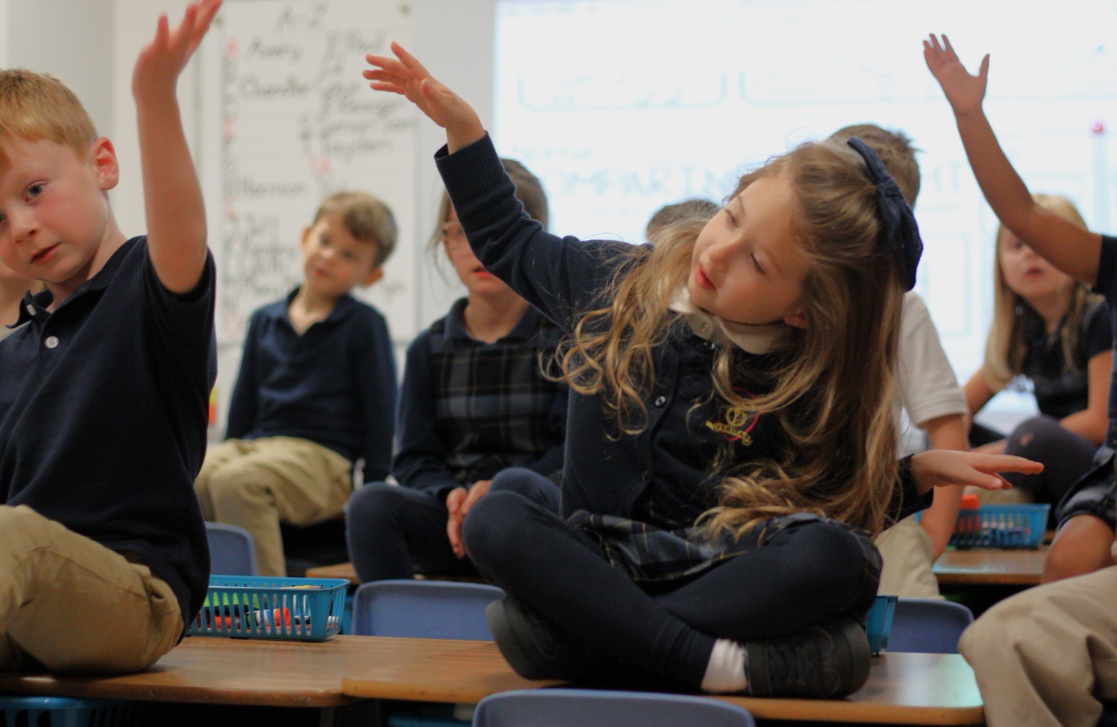 NOCA Lower School students sitting cross-legged on their desks with hands raised, engaged in a classroom lesson.