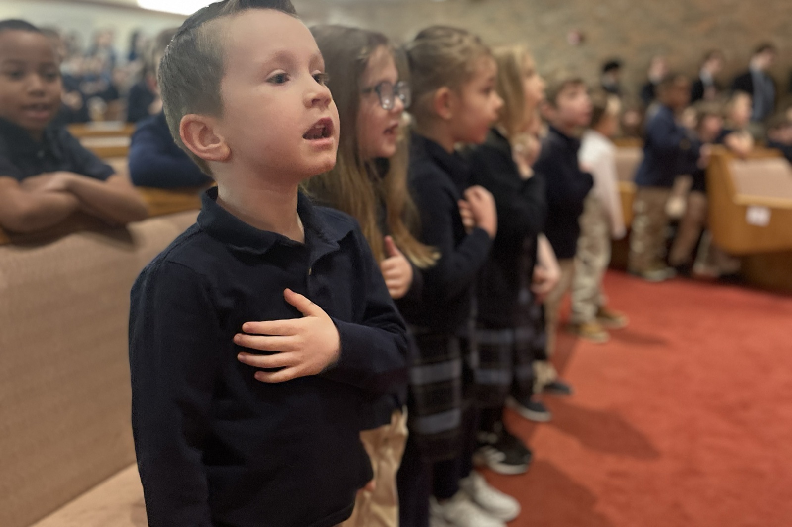A young NOCA Lower School student stands with his hand over his heart during the Pledge of Allegiance at morning assembly, with classmates in a line beside him.